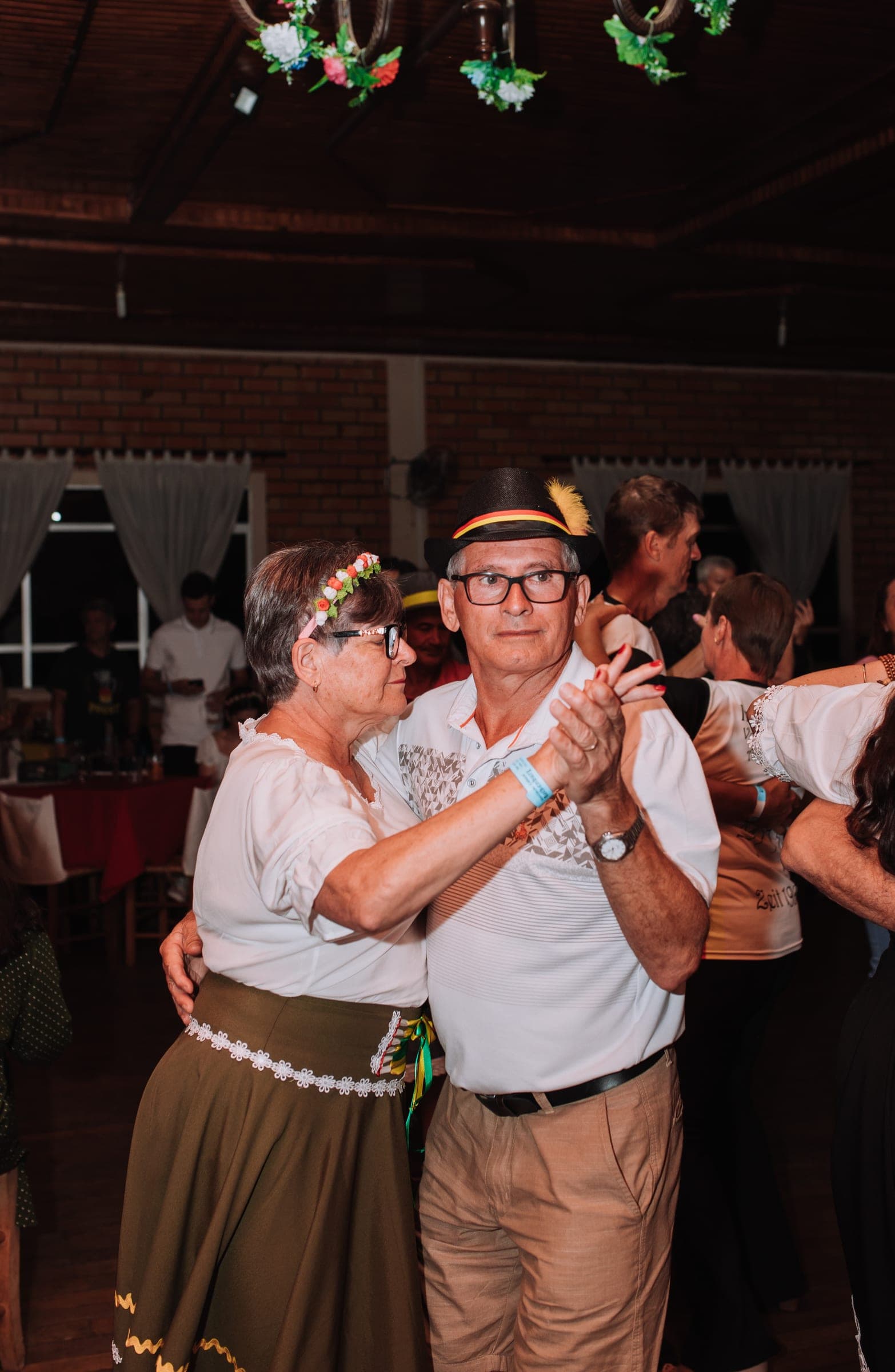 Casal mais velho dançando junto no Baile Alemão, ela de saia tradicional verde-oliva e ele de polo branco com chapéu