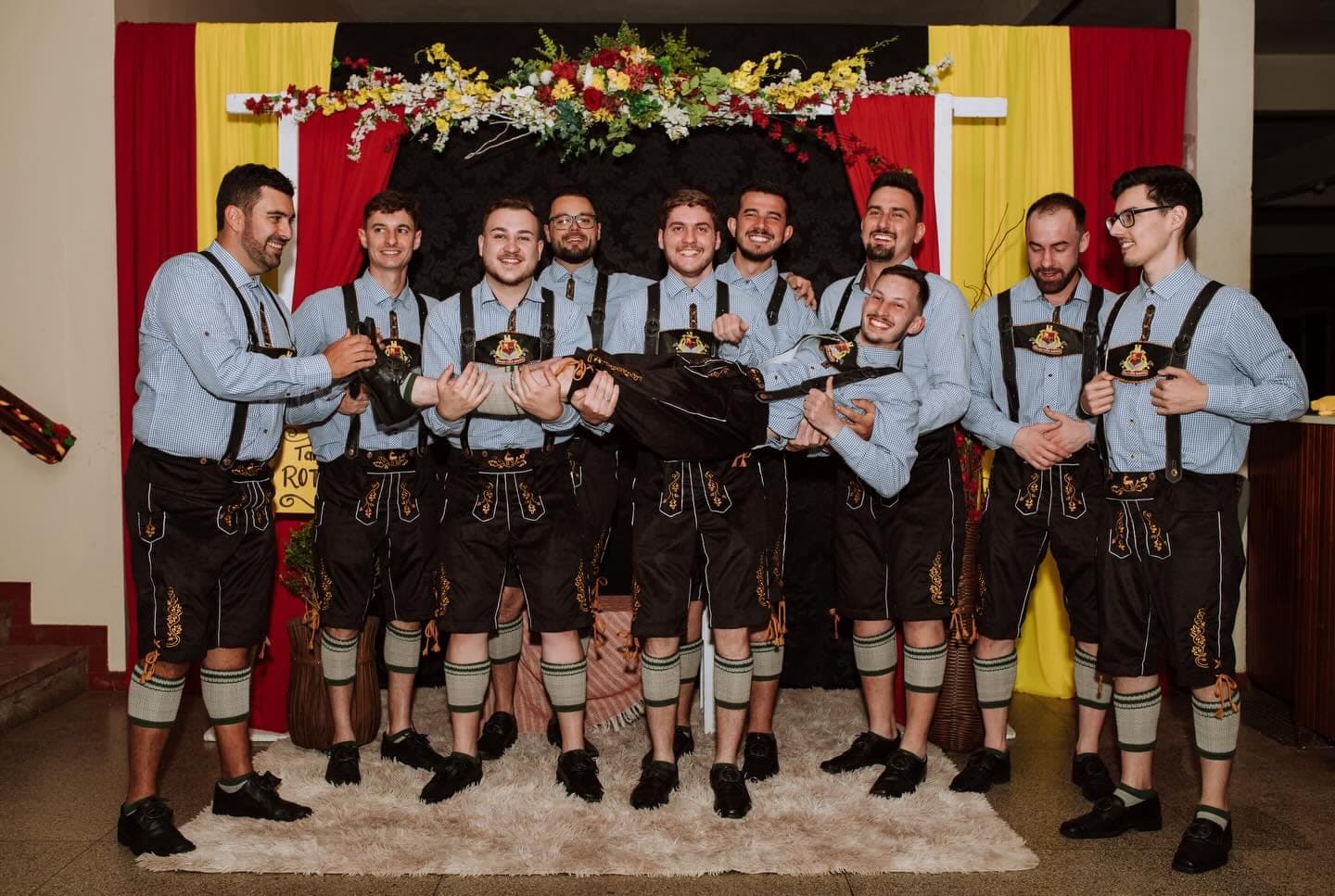 Cavalheiros do grupo de lederhosen segurando um colega no colo, todos rindo